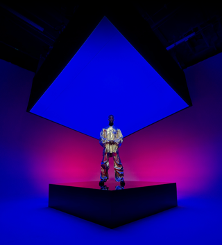 A person dressed in a metallic suit standing on a square center stage and encompassing the background showcasing the Ultra Wide camera on iPhone 16 Pro Max